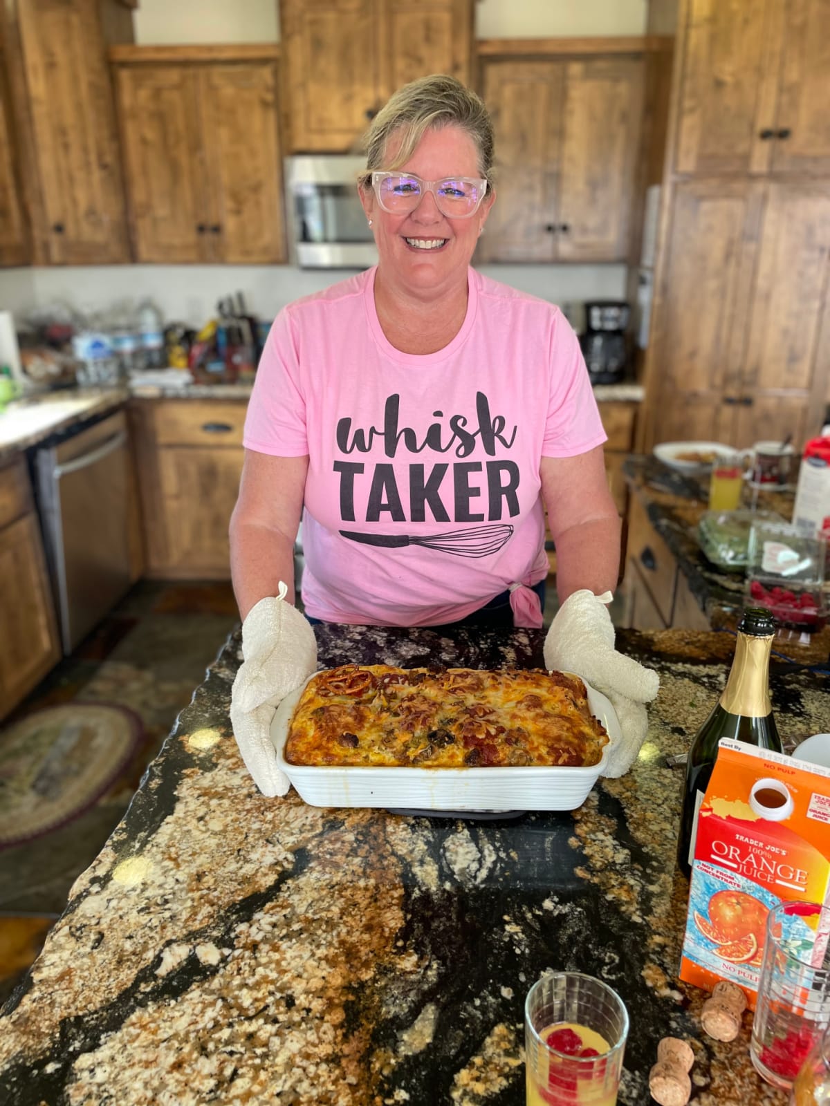 A lady in a kitchen holding a breakfast casserole with oven mitts smiling and about to serve brunch.