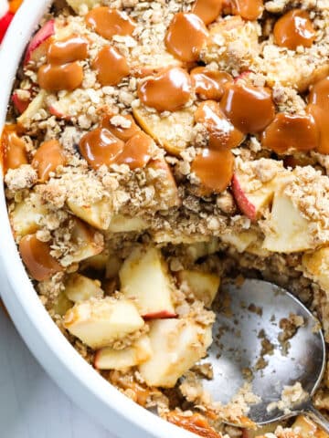 A round white dish of fresh baked caramel apple crisp with a large serving spoon for a Fall dessert.