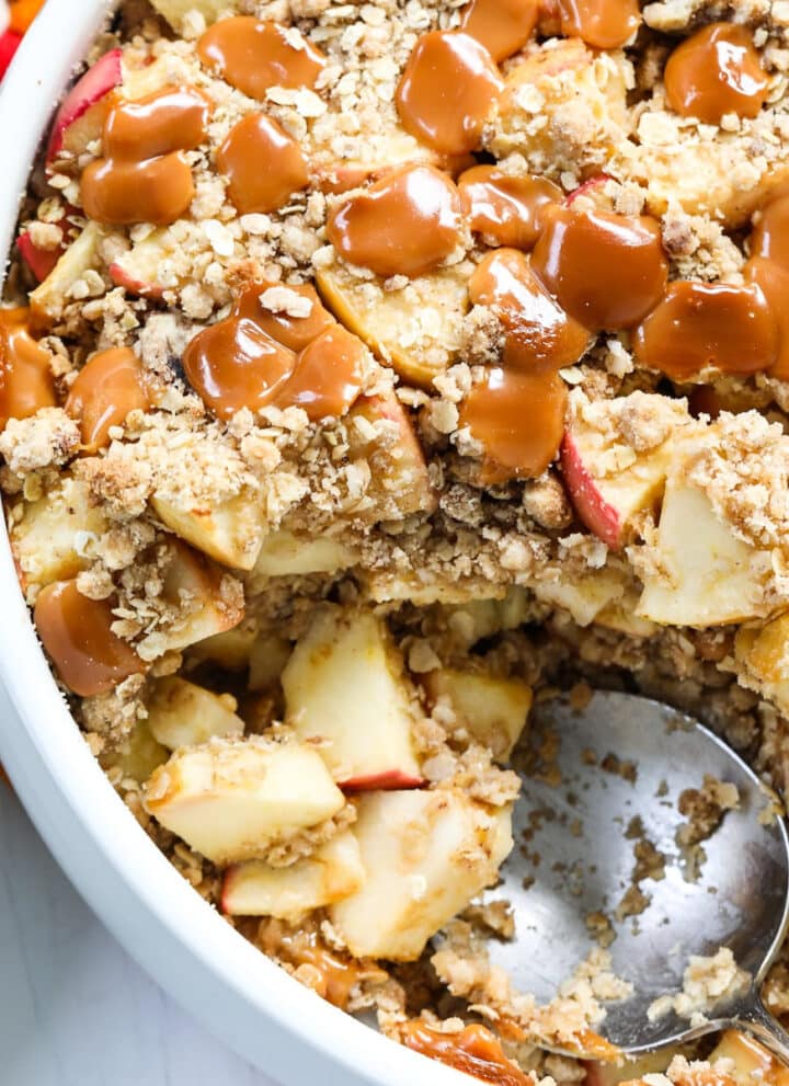 A round white dish of fresh baked caramel apple crisp with a large serving spoon for a Fall dessert.