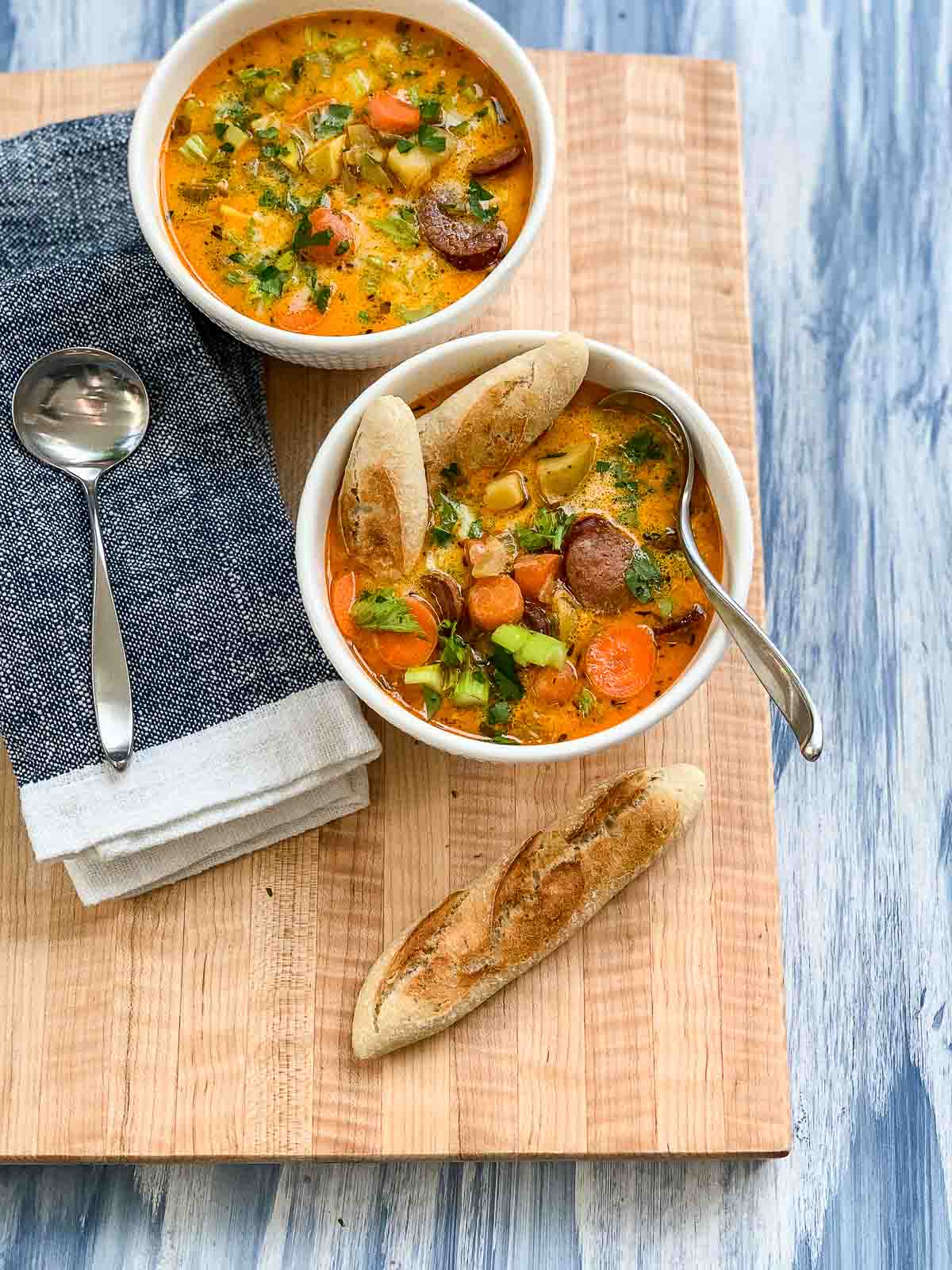 A wooden cutting board with two white bowls of homemad sausage potato soup with soup spoons, a blue napkin, and pieces of bread.