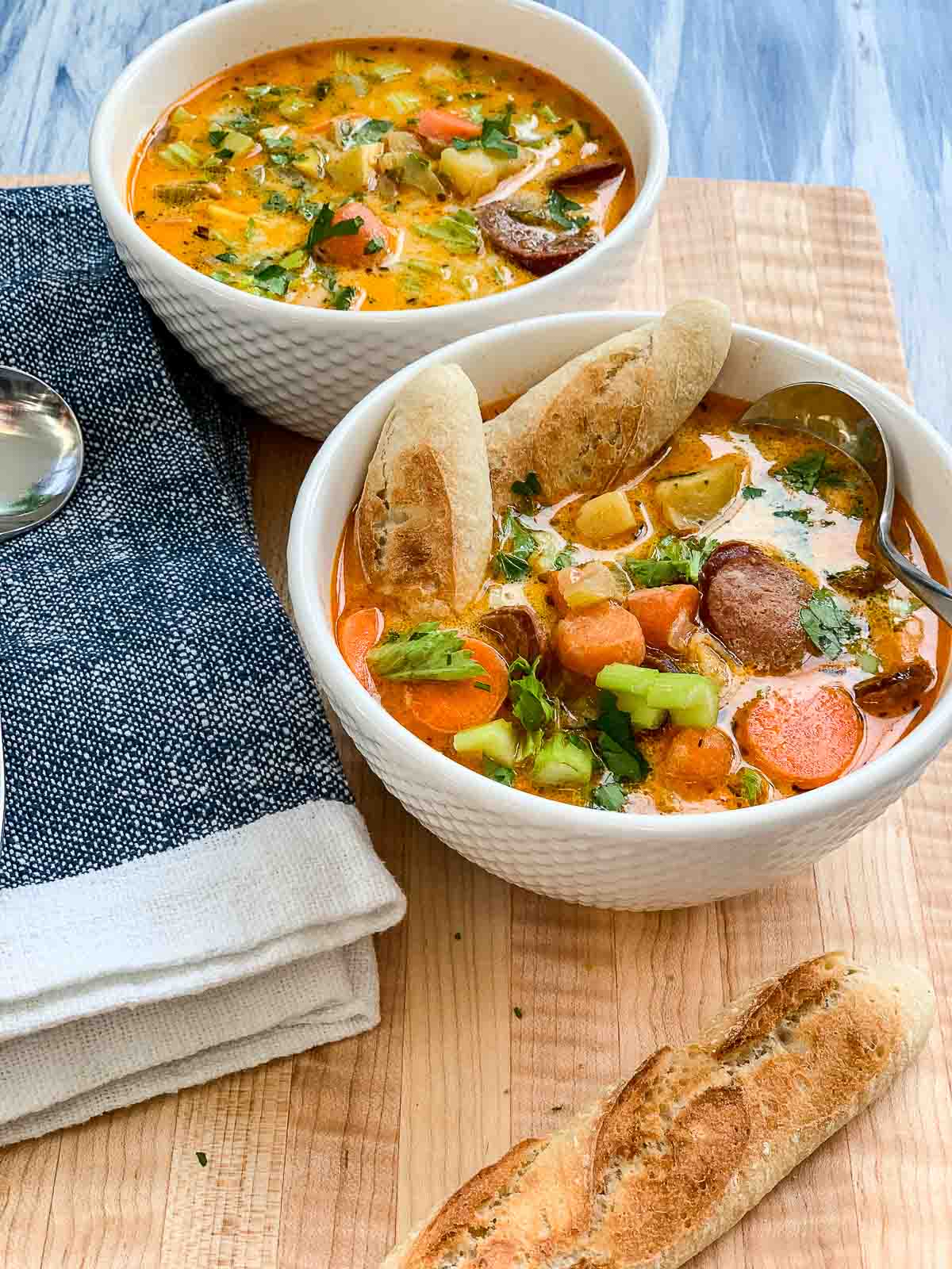 Close up of two white bowls of spicy sausage potato soup with pieces of bread broken inside the soup on a cutting board with a napkin.