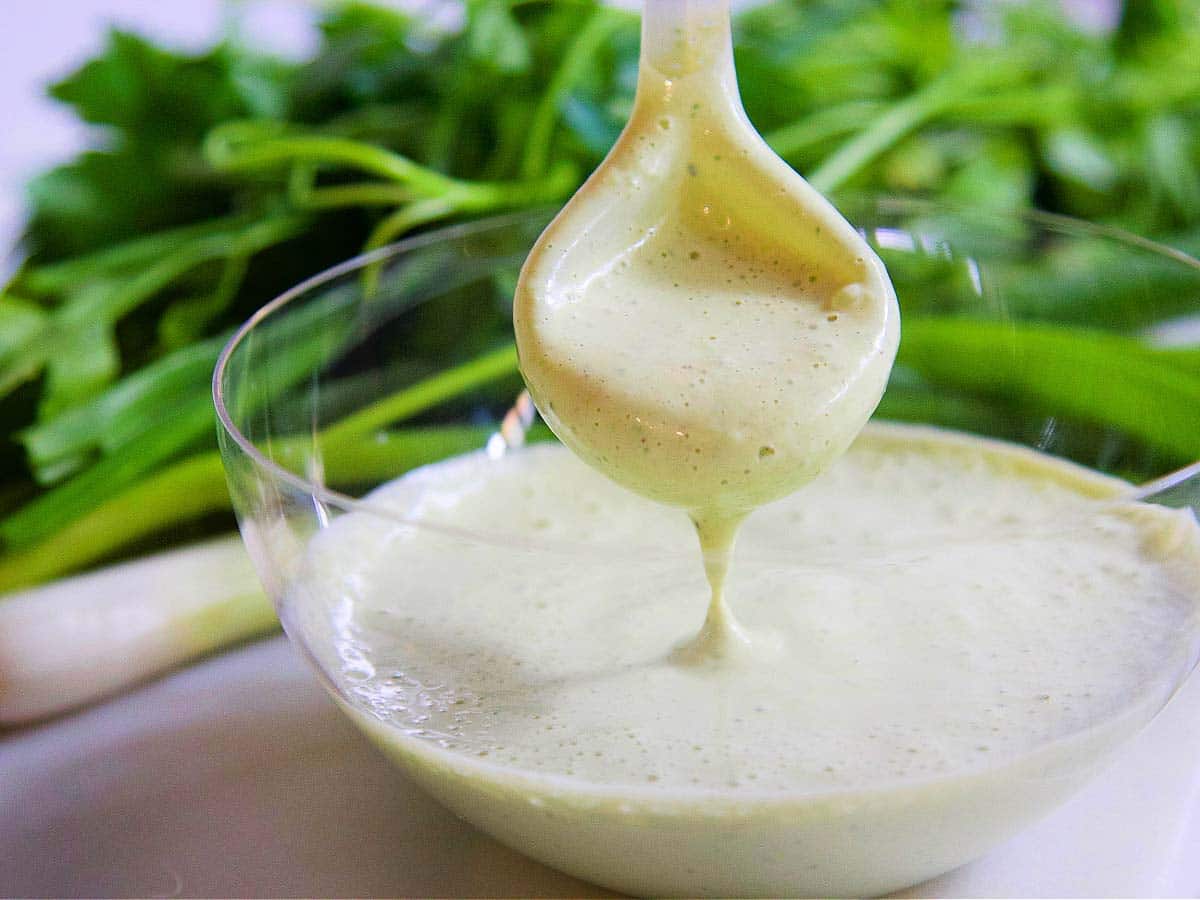 A clear glass bowl with a spoon dipping into Green Goddess dressing. 