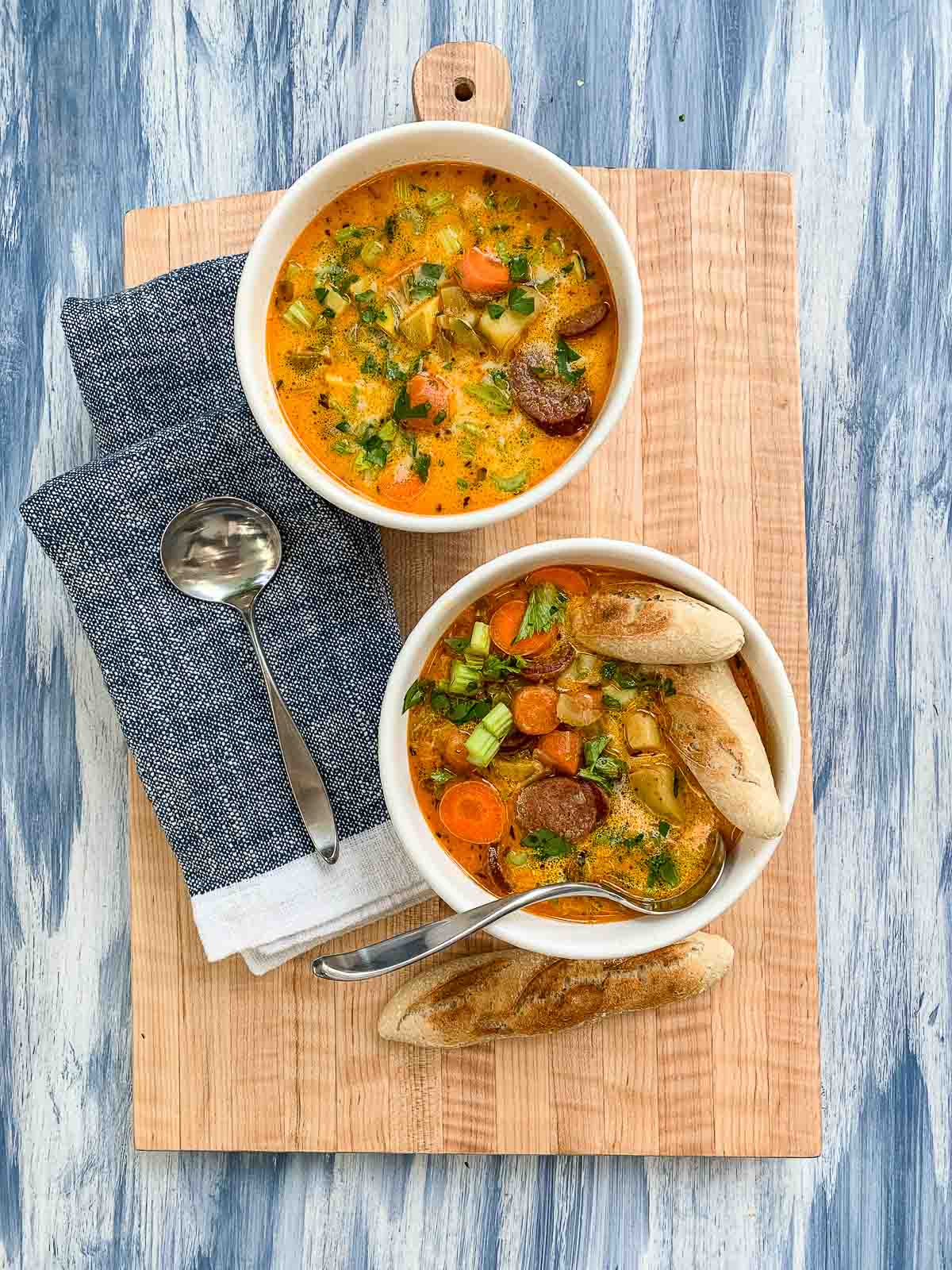 Two white bowls filled with spicy sausage and potato soup with spoons and blue napkins on a cutting board. 