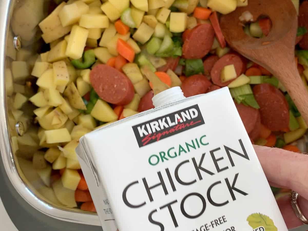 Pouring chicken stock into a slow cooker with chopped vegetables and sausage for a soup recipe.
