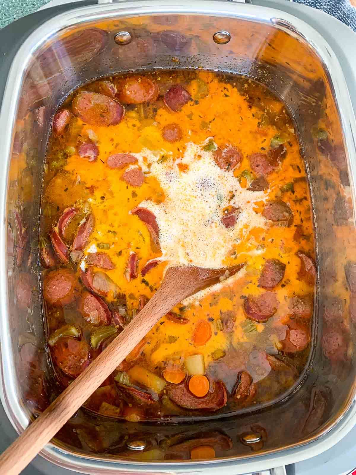 Slow cooker with ingredients inside and heavy cream on top to make soup with a wooden spoon.