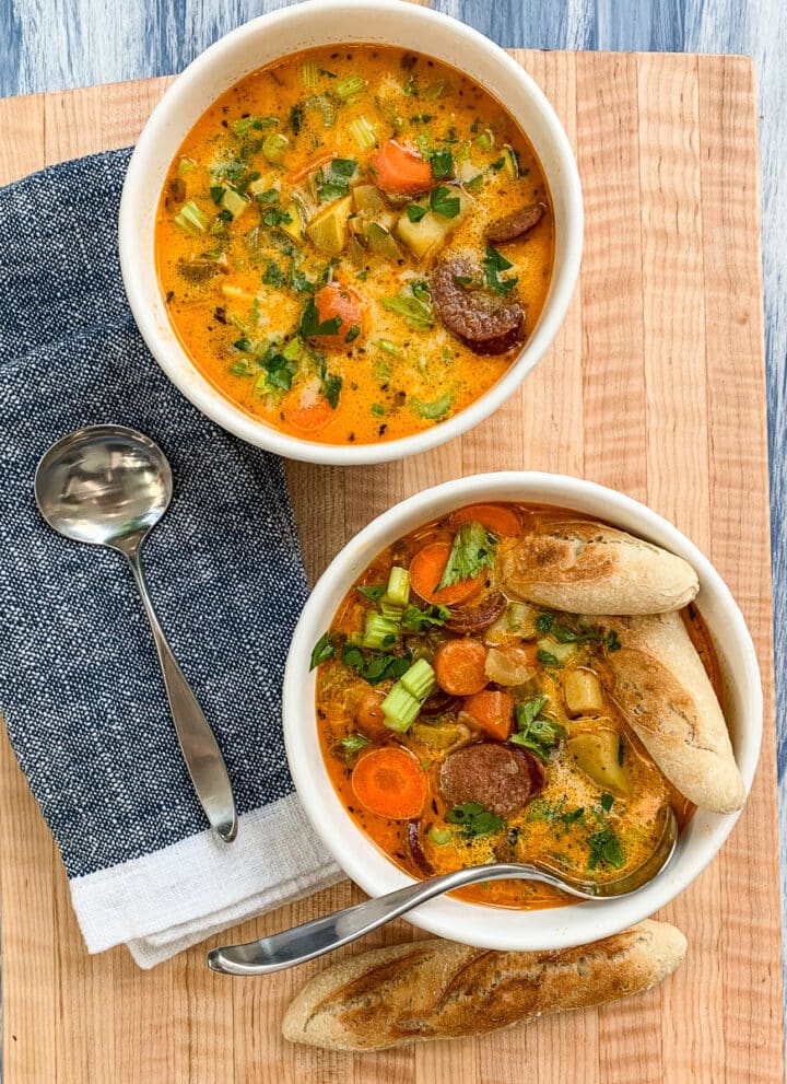 A wooden board with two bowls of sausage potato soup with spoons and pieces of bread.