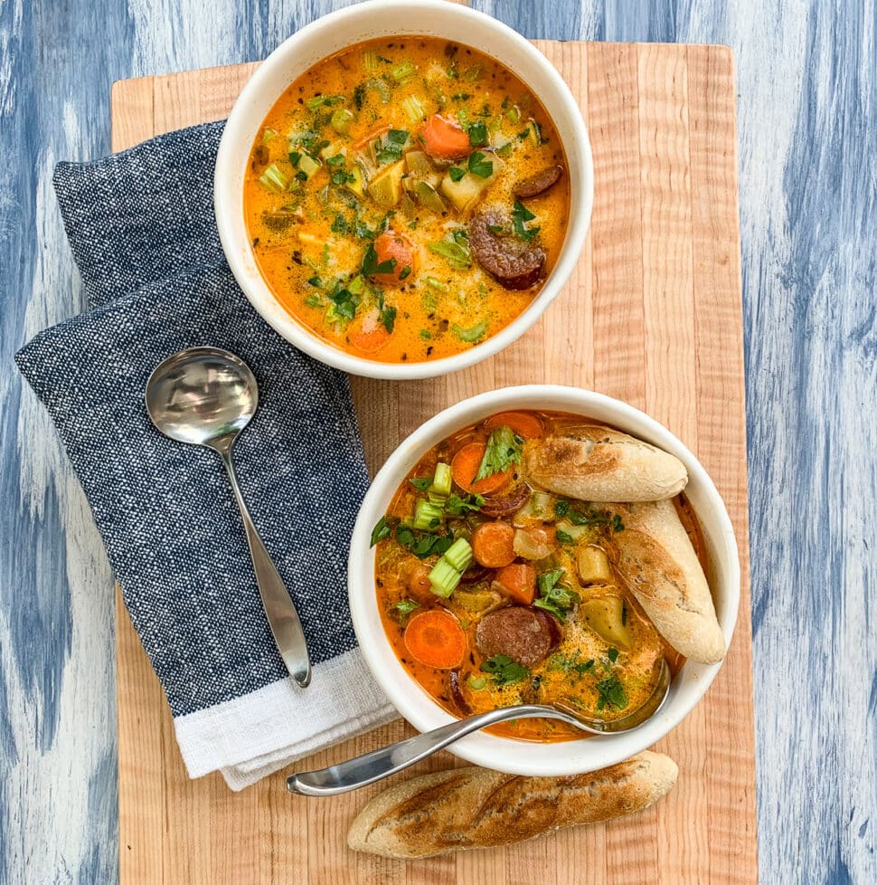 A wooden board with two bowls of sausage potato soup with spoons and pieces of bread.